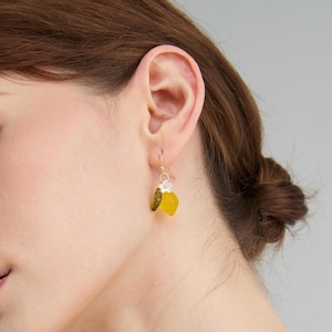 May include: Gold-tone dangle earrings featuring a small white flower and two leaf-shaped charms. One charm is olive green, and the other is a bright yellow. The earrings are designed to be worn with a hook.