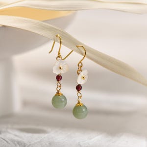 May include: A pair of gold dangle earrings with white flower-shaped shell charms, green jade beads, and small red beads.
