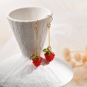 May include: A pair of gold-colored dangle earrings featuring red, textured strawberry charms with green leaf accents. The earrings hang from delicate gold wires, showcasing a playful, fruit-themed design.