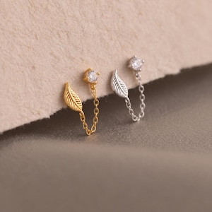 May include: Two double-piercing earrings, one gold-toned and one silver-toned. Each earring features a small round clear gemstone stud connected by a delicate chain to a small leaf-shaped charm. The design allows for two piercings to be linked.