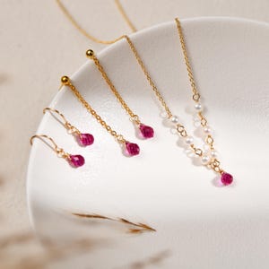 May include: A selection of gold-coloured jewellery, including earrings and necklaces, presented on a white surface. The earrings and one necklace feature teardrop-shaped, vibrant pink gemstones. Another necklace incorporates small white pearls. The jewellery is delicate and elegant.