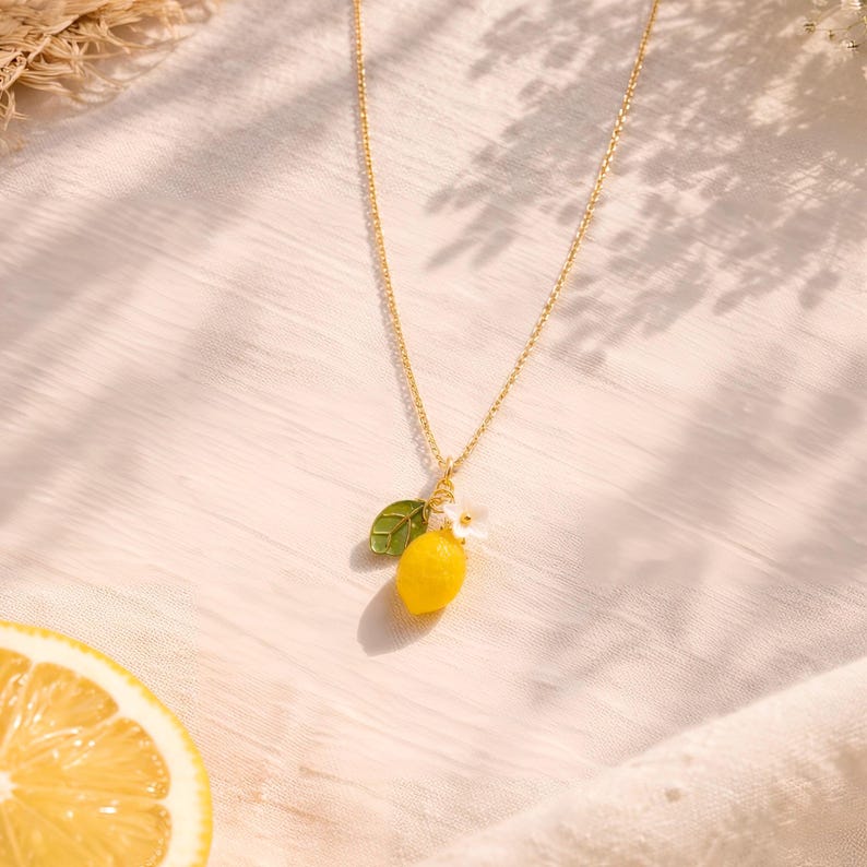 May include: A gold-colored necklace with a pendant featuring a yellow lemon, a green leaf, and a small white flower. The necklace is displayed on a white, textured surface, with a slice of lemon in the lower left corner.