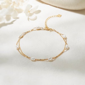 Op de afbeelding: Een delicate goudkleurige enkelband met twee fijne kettingen. Elke ketting is versierd met verschillende ovale, witte parels. De enkelband heeft een verstelbare ketting en een kleine hartvormige bedel.