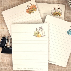 May include: A collection of lined notepads with watercolor pumpkin illustrations in various colors. The pads feature different pumpkin designs, including orange, white, and teal pumpkins, with sunflowers and birds. A black pen and binder clip are visible.