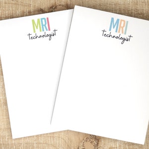 May include: Two white notepad sets with the text "MRI Technologist" in black script and the letters "MRI" in colorful block letters. The notepad on the left has the letters "MRI" in green, pink, and blue. The notepad on the right has the letters "MRI" in blue, orange, and light blue. A black pen is lying on a wooden surface next to the notepads.