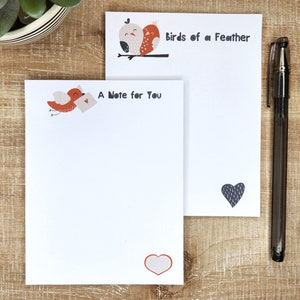 May include: Two white notepads with bird illustrations and text. One notepad reads "A Note for You" with a red bird carrying a letter. The other says "Birds of a Feather." A black pen rests on the side.