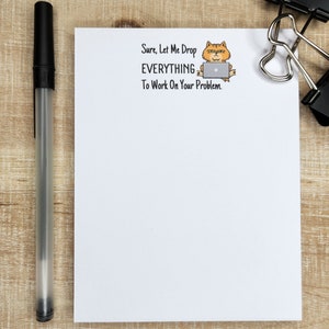 May include: White notepad with a cartoon cat wearing glasses and using a laptop. The text on the notepad reads "Sure, Let Me Drop EVERYTHING To Work On Your Problem."