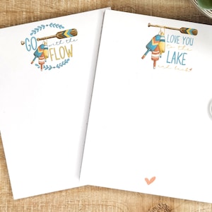 May include: Two blank white notepads with a decorative design on each. The notepad on the left has a blue and yellow oar with the text "GO with the FLOW". The notepad on the right has a blue and yellow oar with the text "LOVE YOU to the LAKE" and a small pink heart.