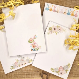 May include: Three white notepads with different Easter-themed designs. One notepad features a floral design with pink and white flowers. Another notepad features a bunny silhouette made of flowers. The third notepad features a bunny with a floral wreath around its neck.