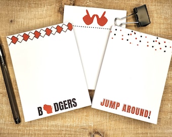 May include: Three white notepads with decorative red and black accents. One notepad has the text "BADGERS" with a red Wisconsin outline. Another says "JUMP AROUND!" in red. A black pen and binder clip are also present.