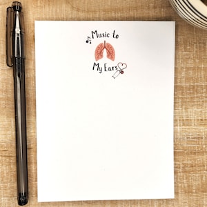 May include: A white notepad with the phrase "Music to My Ears" and a graphic of lungs, a musical note, and a stethoscope. A black pen rests on the left side of the notepad. The notepad is on a wooden surface.