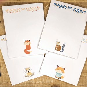 May include: Four white notepads with illustrated foxes in various poses. Each notepad has a different fox design, with orange, brown, and blue accents. Decorative floral elements are at the top of each notepad. A black pen is visible on the left.