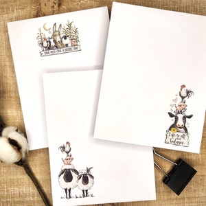 May include: Three white notepads with different farm animal illustrations. The top notepad features a cow, pig, rooster, and rabbit with the text "Cluck Moo Cock & Doodle Doo". The middle notepad features two sheep with a rooster and pig on top of one of the sheep. The bottom notepad features a cow with a pig and rooster on top of its head. The text on the bottom notepad says "Life is all about balance".