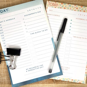 May include: Two daily planner notepads with "TODAY" at the top. One notepad has a blue and white design, the other has a floral border. A black pen rests on the floral notepad. A black binder clip holds the blue notepad.