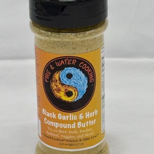 May include: A glass jar with a black lid filled with a yellow powder. The label reads "Fire & Water Cooking" with a yin yang symbol and "Black Garlic & Herb Compound Butter". The label also says "Try on Beef, Pork, Poultry, Seafood, Veggies, and More!" and "Hand Crafted**Gluten & MSG Free**" with a net weight of 8.7 oz.