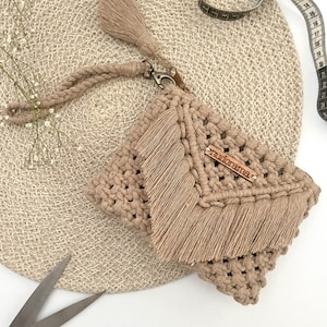 Op de afbeelding: Een beige gehaakte clutch met een franje detail en een klein metalen label met de tekst "remarkable".
