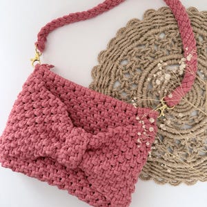 Puede incluir: Un bolso de mano de macramé en color rosa empolvado con un patrón de nudos texturizados en la parte frontal y un detalle de lazo central. El bolso incluye una correa trenzada para el hombro sujeta con cierres de mosquetón de metal dorado.