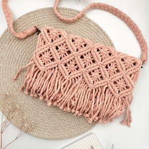 May include: A pink macrame crossbody bag with a fringe detail. The bag is sitting on a woven round mat.
