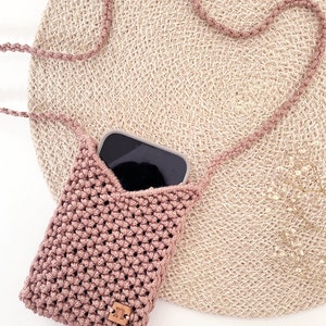 Könnte beinhalten: Eine kleine, gehäkelte rosa Handytasche mit einem braunen Lederanhänger. Die Tasche liegt auf einer geflochtenen Strohmatte.