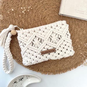 Op de afbeelding: Crèmekleurige macramé clutch met een geometrisch patroon en een polsbandje. De clutch heeft een houten label met de tekst "nudorama". Een kwastje en houten kraal accentueren de band. De clutch ligt op een geweven bruine mat.