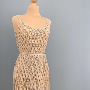 Könnte beinhalten: Beiges Makramee-Kleid auf einer Schaufensterpuppe. Das Kleid hat ein rautenförmiges Muster, einen Rundhalsausschnitt und dünne Träger. Ein passender Gürtel schnürt die Taille. Das Kleid ist aus Naturfaser gefertigt und reicht bis zur Mitte der Wade.