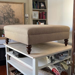 Peut inclure: Un pouf carré recouvert d'un tissu pied-de-poule marron et beige. La pièce présente un dessus plat et rembourré et repose sur quatre pieds en bois tourné de couleur marron foncé. Le design est simple, avec des lignes épurées.
