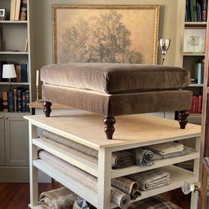 May include: A rectangular taupe velvet ottoman featuring a thick cushioned top and four dark brown turned wooden legs. The ottoman is positioned on a white wooden table with multiple open shelves containing various rolled fabric textiles.