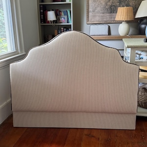 May include: A freestanding upholstered headboard featuring a curved, arched top silhouette. The fabric is a light beige and white vertical pinstripe pattern, finished with a dark brown piping trim along the edges of the arched frame.