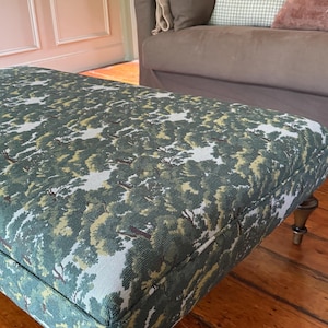 Peut inclure: Un pouf rectangulaire recouvert d'un tissu tissé présentant un motif de paysage forestier dense dans des tons de vert foncé, olive et gris clair. Le meuble est soutenu par quatre pieds en bois tourné brun foncé avec une finition polie.