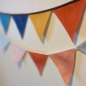 May include: A string of fabric pennants in various colors, including orange, yellow, blue, pink, and green. The pennants are strung together with white string and are hanging on a white wall.