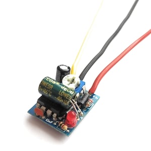 May include: A blue circuit board with electronic components, including a capacitor, resistor, and LED. The board has the text "CON OWESR 0+105°C -40+105" and "JEGO LOWES" printed on it. Three wires are connected to the board, one red, one black, and one yellow.