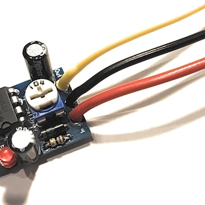 Può includere: Una scheda di circuito blu con un piccolo chip nero, una manopola bianca e tre fili collegati ad essa. I fili sono rosso, nero e giallo. La scheda ha il testo "Set Up" e "Out" stampato su di essa.