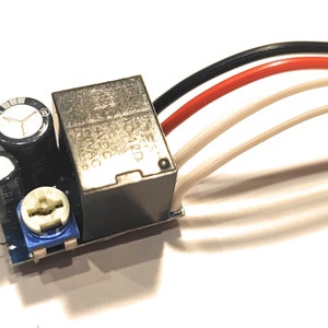 Op de afbeelding: Een klein elektronisch circuitbord met een zwarte relais, twee condensatoren, een blauwe potentiometer en drie draden die eraan zijn verbonden. De draden zijn zwart, rood en wit.