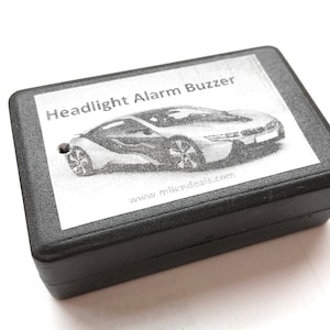 Puede incluir: Una caja de plástico negra con el texto "Headlight Alarm Buzzer" y una imagen en blanco y negro de un coche. La dirección del sitio web "www.infideals.com" está impresa en la caja.