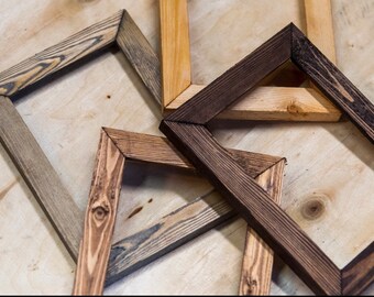 May include: Four wooden picture frames in different shades of brown, gray, and black. The frames are made of natural wood with visible grain patterns. They are arranged in a pile on a light wood surface.