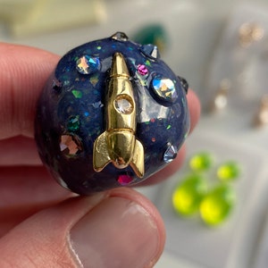 May include: A dark blue resin ring with a gold rocket ship design and sparkly rhinestones. The ring is decorated with a variety of colorful rhinestones.