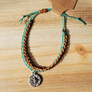 May include: A turquoise and brown braided bracelet with a silver compass charm.