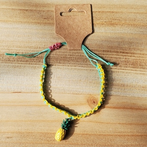 May include: A yellow and green pineapple charm bracelet with a brown tag. The bracelet is made of braided string.