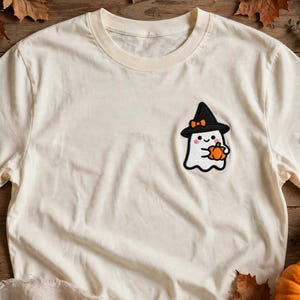 May include: A cream-colored short-sleeve t-shirt featuring a small embroidered graphic on the left chest. The graphic depicts a white ghost wearing a black witch hat with an orange bow, holding a small orange pumpkin with a green stem.