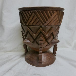 May include: A handcrafted wooden container with a lid, featuring intricate geometric carvings. The container is a warm brown color and stands on a pedestal base with decorative legs. The design includes chevron and angled patterns.