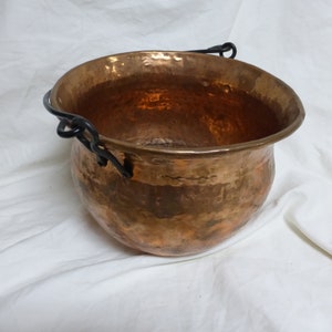 May include: A hammered copper cauldron with a rounded body and a flared rim. The cauldron has a dark, wrought iron handle. The copper has a warm, reddish-brown color and a textured surface, suggesting it is handmade.