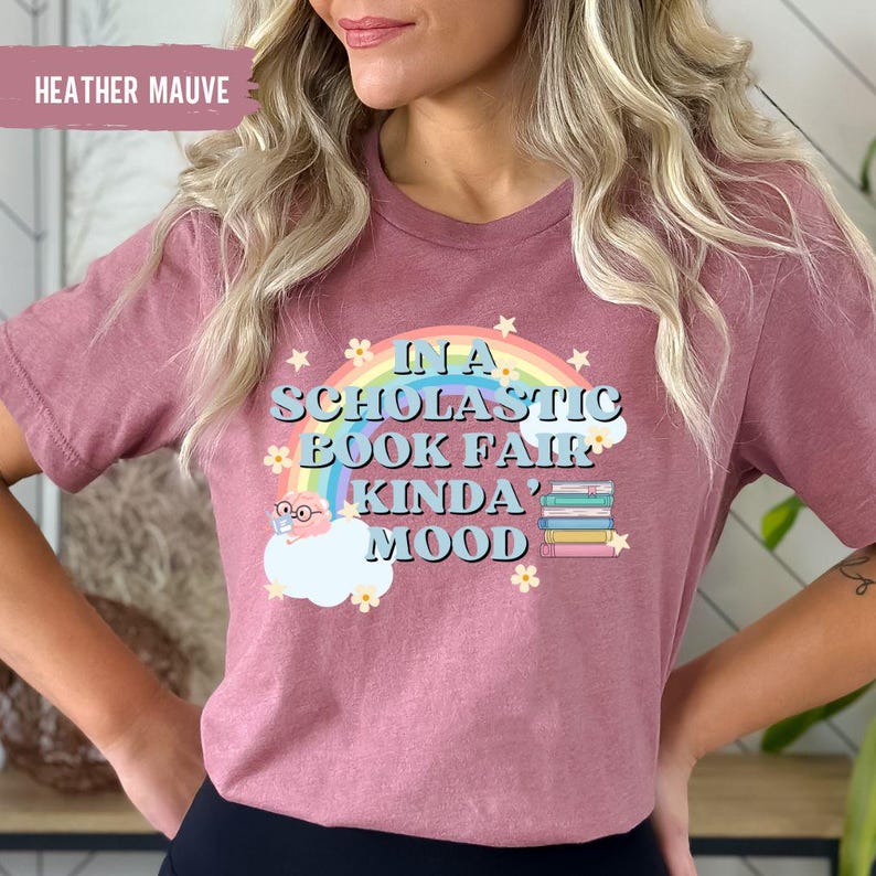 Puede incluir: Camiseta de color malva heather con un arco&iacute;ris, estrellas y un gr&aacute;fico de nube. El texto de la camiseta dice "In a Scholastic Book Fair Kinda' Mood".