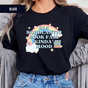 Puede incluir: Camiseta negra con un gr&aacute;fico de arco&iacute;ris y nubes. El texto "In a Scholastic Book Fair Kinda' Mood" est&aacute; impreso en la camiseta.