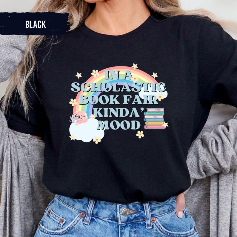 Puede incluir: Camiseta negra con un gr&aacute;fico de arco&iacute;ris y nubes. El texto "In a Scholastic Book Fair Kinda' Mood" est&aacute; impreso en la camiseta.