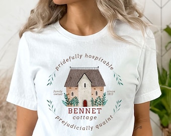 May include: White t-shirt featuring a watercolor illustration of a cottage with the words "Bennet Cottage" and surrounding text. The design includes the phrases "pridefully hospitable" and "prejudicially quaint."