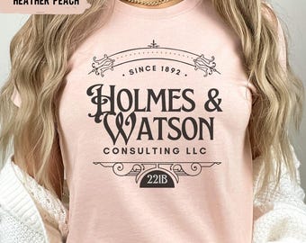 May include: Heather peach-colored t-shirt with the text "HOLMES & WATSON CONSULTING LLC" in black, along with "SINCE 1892" and the address "221B". The shirt has a crew neck and short sleeves.