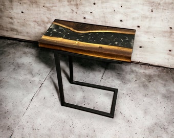 May include: A wooden side table with a black epoxy resin top featuring a river design with black stones. The table has a black metal frame.