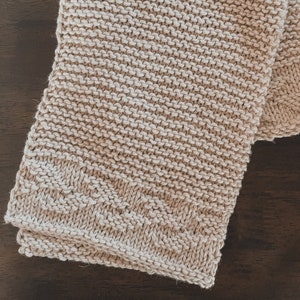 May include: A beige knitted dishcloth with a textured pattern. The dishcloth is folded in half and is lying on a wooden surface.