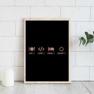 以下が含まれることがあります： 「eat(); code(); sleep(); repeat();」という言葉が銅色で書かれた、額入りの黒いプリント。明るい木製のフレームで、白いタイルの背景に表示されています。2本の白いろうそくが前景にあります。