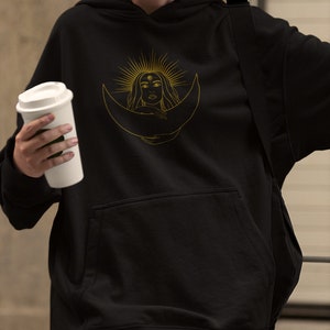 Könnte beinhalten: Schwarzer Kapuzenpullover mit einer goldenen Illustration einer Frau in einem Halbmond, umgeben von Sonnenstrahlen. Der Hoodie hat eine Vordertasche und eine Kapuze mit Kordelzug. Das Design ist auf der Brust zentriert.